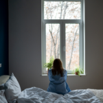 Das Bild zeigt eine Frau, die mit dem Rücken zum Betrachter auf einem Bett sitzt. Sie schaut aus dem Fenster, und ihre Haltung vermittelt Traurigkeit und Nachdenklichkeit.